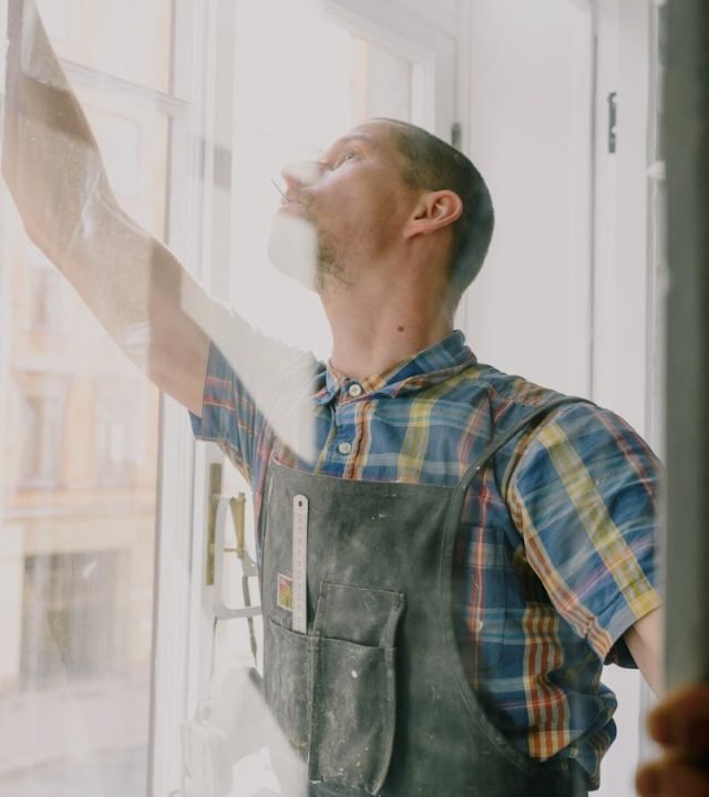 Professional focused craftsman in uniform standing with raised arm standing behind glass while assembling window in apartment during renovation works